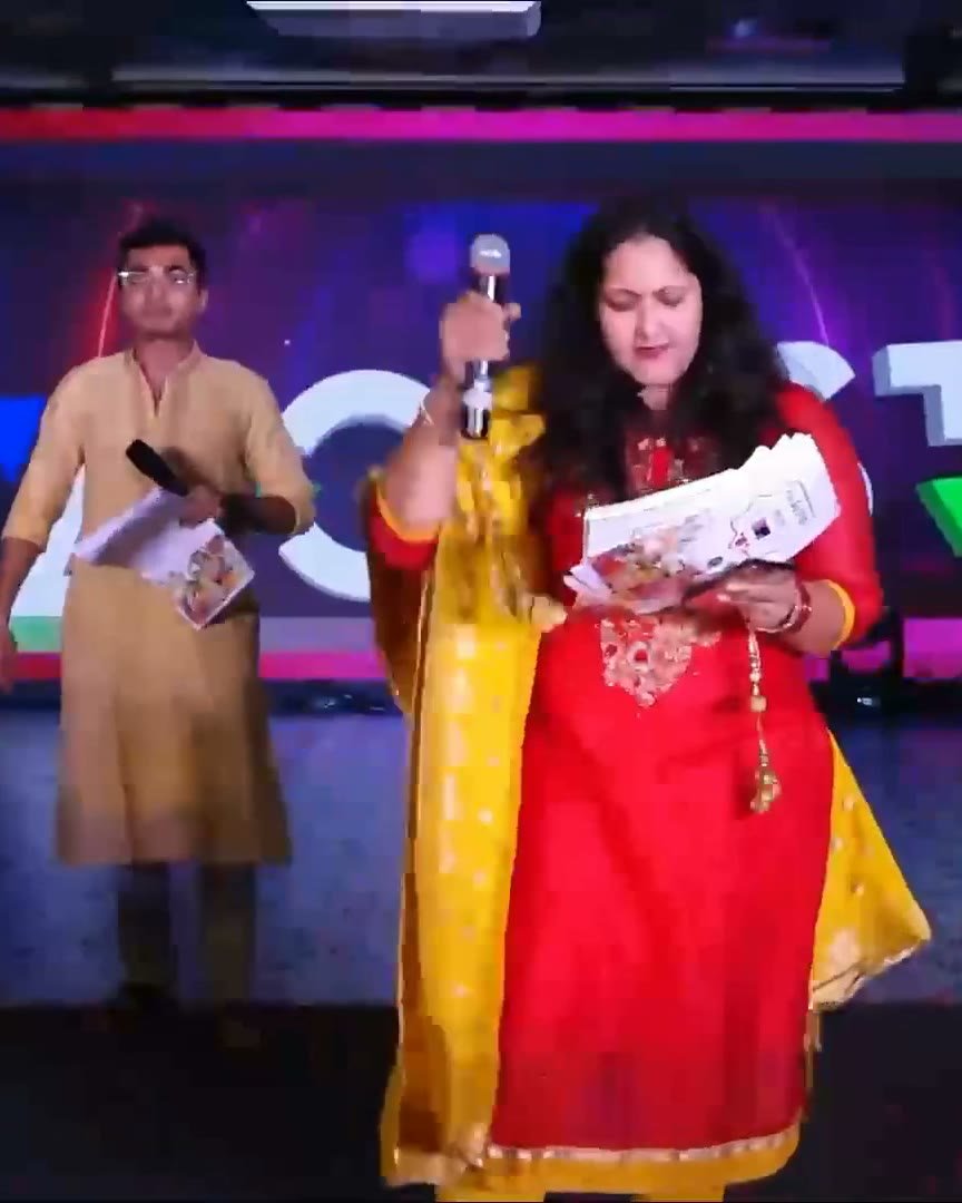 Pooja Aravalapalli on stage with a co-host, microphone raised, in front of a brightly lit LED wall
