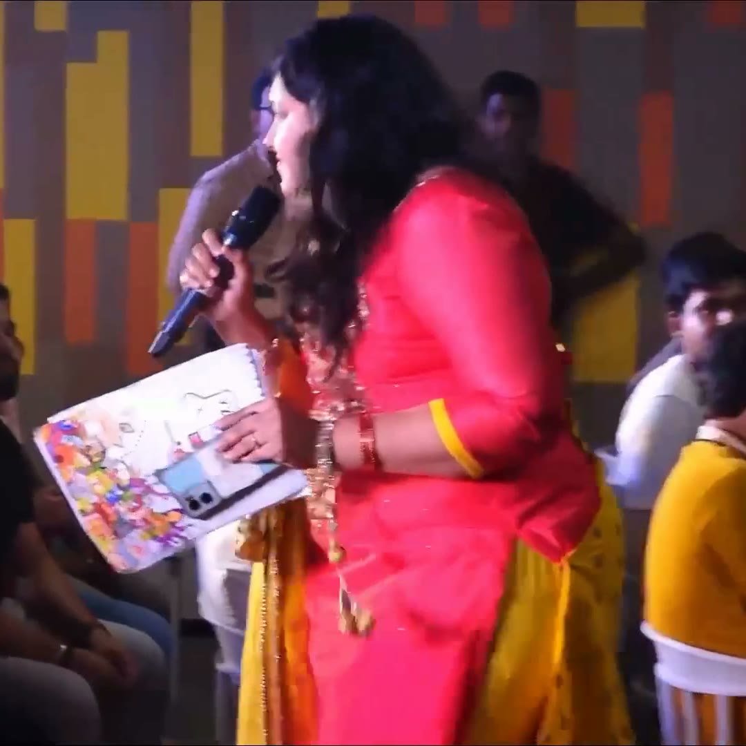 Pooja Aravalapalli standing among the seated audience with a microphone and run sheet, addressing the room