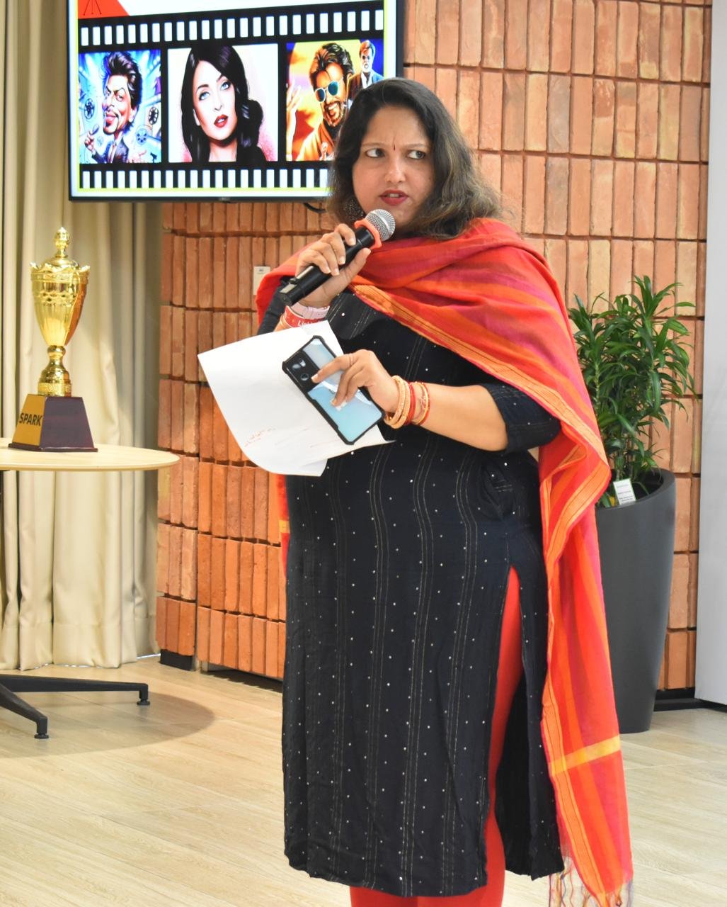 Pooja Aravalapalli hosting a corporate event, microphone in hand, with an award trophy on the table beside her
